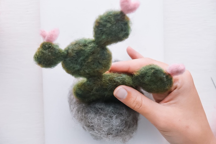 Bovenaanzicht van een witte tafel met daarop een gevilte wollen cactus met roze bloemetjes.