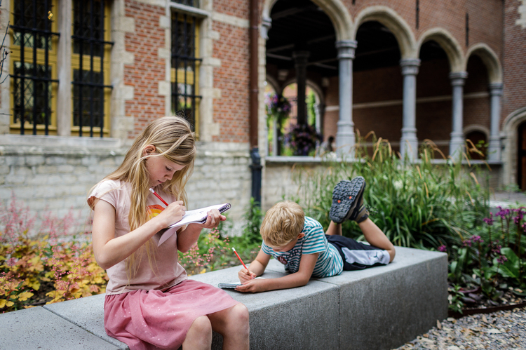 Two children are sitting on a bench in the museum garden. They are writing or drawing in a notebook.