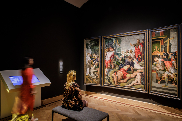 Twee bezoekers kijken naar een groot drieluik schilderij.