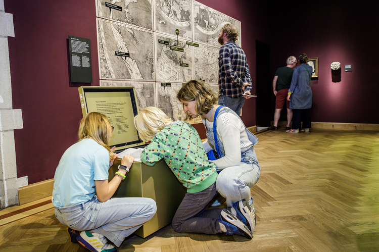 Two children and an adult are looking into a golden cube that's sitting in a museum hall.