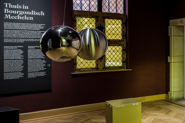 Twee grote metalen bollen hangen in een donkerrode ruimte. Één van de bollen is glanzend en reflecteert de ruimte, de andere is mat en wazig.